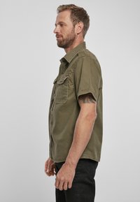 Cămașă scurtă, de culoare verde măslin, cu guler tip button-down, două buzunare pe piept și o croială casual, prezentată pe un bărbat din lateral.