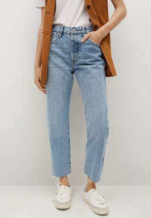 Person iført lyseblå højtaljede cropped jeans med frynser i kanten, hvide sneakers, hvid t-shirt og brun ærmeløs jakke.