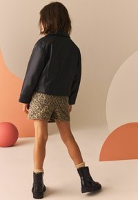 Zwarte nepleer jas met een relaxte pasvorm, gecombineerd met beige shorts met luipaardprint en zwarte enkellaarsjes met geribde crèmekleurige sokken.