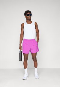 Sportlicher Mann in einem weißen Tanktop, leuchtend pinken Shorts, Sportsonnenbrille und weißen Sneakers, der in der rechten Hand eine schwarze Wasserflasche hält.