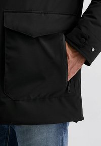 Zwarte waterdichte jas met een grote voorzak, ritsdetails en knoopaccenten op de manchetten. Glad textuur, modern ontwerp.