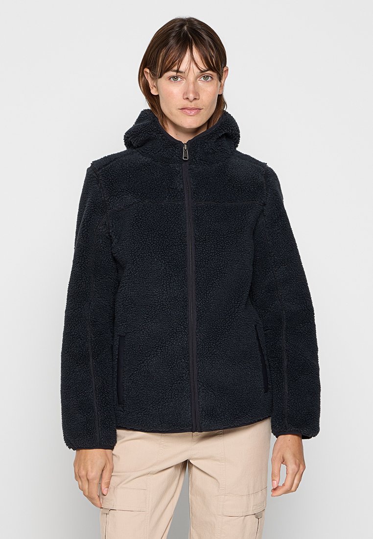 Femme portant une veste en polaire noire zippée avec capuche et un pantalon cargo beige, se tenant devant un fond clair.