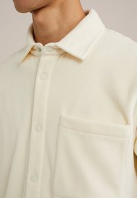 Cream fleece shirt featuring a front pocket, collared neckline, and snap closures. Soft texture with a smooth finish.