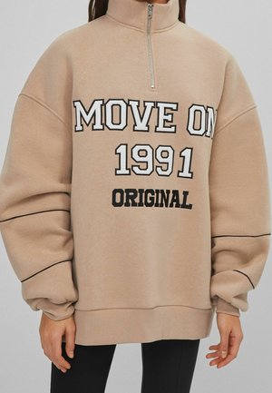 Beżowa bluza z suwakiem do ćwiartki, z bufkami na rękawach, czarną lamówką i napisem "MOVE ON 1991 ORIGINAL" pogrubionymi białymi i czarnymi literami.