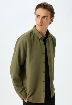 Camisa de botones en verde olivo, de tejido texturizado, con un cuello clásico y mangas largas, llevada sobre una camisa negra.