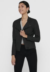 Zwarte neplederen jas met ribbelstructuur op de schouders en mouwen, ritszakken en een asymmetrische sluiting aan de voorkant.