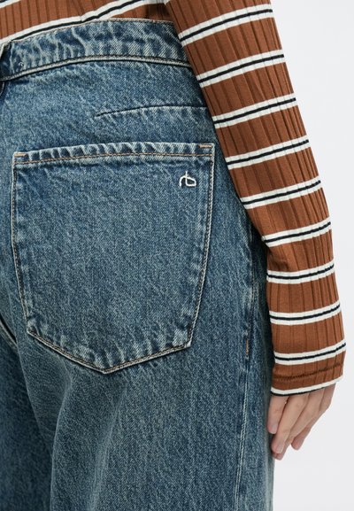 Calças de ganga azul de cintura subida com acabamento texturizado, apresentando um bolso traseiro detalhado e costuras subtis, combinadas com um top de malha canelada às riscas castanho.