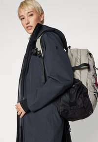 Black waterproof jacket with a high collar, paired with a grey and black backpack featuring mesh pockets and adjustable straps.