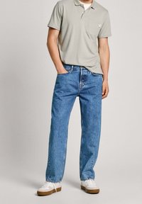 Polo de color gris claro con mangas cortas, detalle de bolsillo, combinado con unos vaqueros de denim azul de corte holgado y zapatillas blancas con detalles en tono marrón.