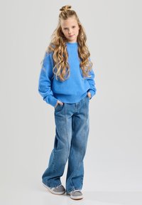 Pull en maille bleu clair, jeans en denim coupe ample, baskets grises avec semelle blanche. Cheveux longs et ondulés, mains dans les poches, se tient contre un fond neutre.