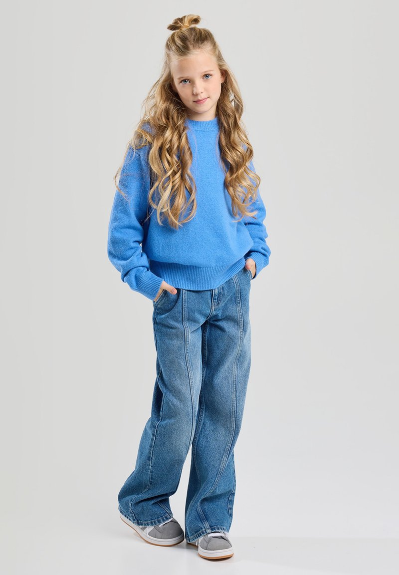 Pull en maille bleu clair, jeans en denim coupe ample, baskets grises avec semelle blanche. Cheveux longs et ondulés, mains dans les poches, se tient contre un fond neutre.