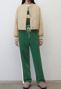 Giacca bomber beige trapuntata, pantaloni verdi con righe laterali, maglione a righe multicolore in maglia e scarpe slip-on viola con foderatura in pelliccia.