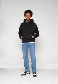 Black hoodie with front pocket and logo, paired with light blue jeans. Black and white sneakers. Smooth cotton fabric, relaxed fit.