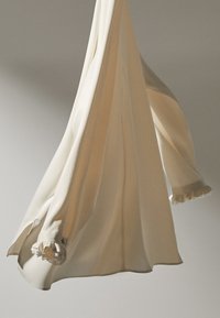 Vêtement en tissu crème avec un design fluide, présentant des accents froncés au niveau des poignets, suspendu contre un fond neutre.