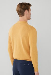 Pullover giallo a maglia con maniche lunghe e colletto polo. Texture morbida, design aderente e dettagli a costine sul bordo. Indossato con pantaloni blu.