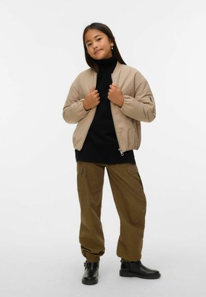 Giovane che indossa una giacca bomber beige sopra un dolcevita nero, pantaloni cargo marroni e stivaletti neri su uno sfondo bianco uniforme.