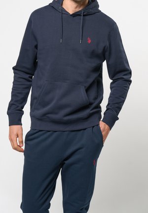 Mann trägt dunkelblauen Kapuzenpullover und passende Jogginghose mit roten, auf Brust und Oberschenkel gestickten Logos, steht mit einer Hand in der Tasche.