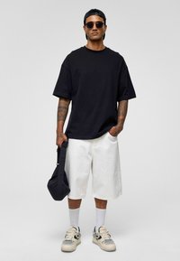 Zwart T-shirt met korte mouwen, witte knielange shorts, witte sokken, beige sneakers, met een zwarte tas.