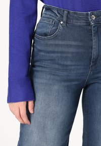 Hoch geschnittene, dunkelblaue Jeans aus Denim mit einer glatten Textur, fünf Taschen und einem Metallknopfverschluss, kombiniert mit einem lila Strickpulloverärmel.