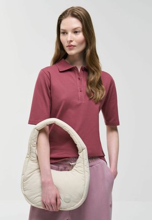 Vrouw met lang bruin haar, gekleed in een mauve poloshirt en roze broek, die een crèmekleurige gewatteerde handtas vasthoudt tegen een lichte achtergrond.
