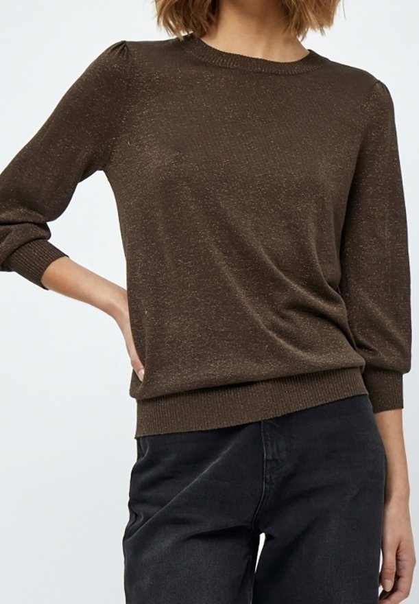 Femme portant un pull marron à manches longues avec des épaules légèrement bouffantes et un pantalon noir, posant avec une main sur la hanche.