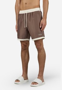 Barna fürdőshorts fehér díszítéssel, elasztikus derékrésszel és húzózsinórral. Kivarrt logó található a lábszáron. Fehér papucsokkal viselve.