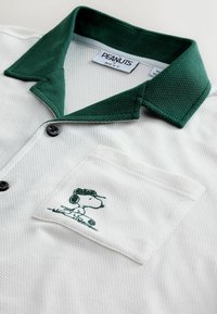 Hvid tekstureret polo med mørkegrøn krave og broderet Snoopy-figur på brystlommen, mærket "Peanuts by Next."