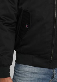 Veste bomber noire avec fermeture éclair à l'avant, poignets côtelés et poche latérale avec un accent logo rond. Texture de tissu lisse.