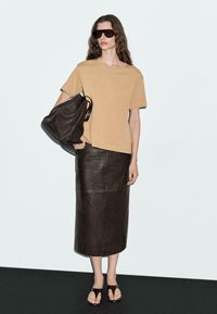 T-shirt in cotone beige, gonna midi in pelle scura, occhiali da sole oversize neri, borsa a mano in pelle marrone e sandali piatti marroni. Design semplice.