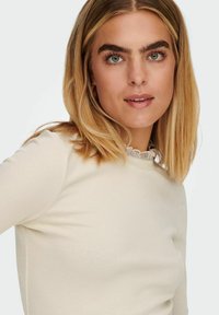 Narmaste soonteedega kreemivärvi pikkade varrukatega pluus, millel on laineline kaelus. Sile tekstuur ja tihe disain rõhutavad rõiva vormi.