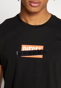 Camiseta de algodón negra con un gráfico rectangular naranja que presenta el texto "DIESEL" y una franja negra brillante a través del diseño.