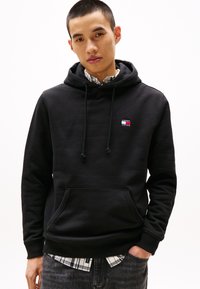 Schwarze Kapuzenjacke aus weichem Stoff, mit Kordelzügen, einer Fronttasche und einem kleinen, farbigen Markenlogo auf der Brust. Über einem karierten Hemd getragen.