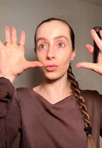Kvinde med flettet hår holder hænder op nær sit ansigt med spredte fingre, iført en brun top mod en ensfarvet baggrund.