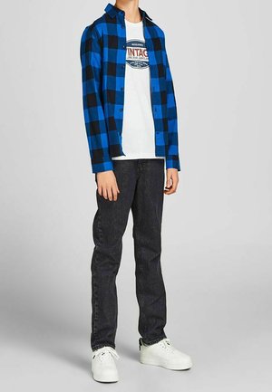 Person trägt ein blau-schwarz kariertes Hemd über einem weißen T-Shirt mit Grafik, schwarze Jeans und weiße Sneaker und steht vor einem grauen Hintergrund.