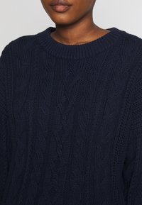 Merensininen neulottu pusero, jossa on resorireunainen pyöreä kaula-aukko, letinmuotoisia kuvioita ja teksturoitua yksityiskohtaisuutta koko kankaassa.