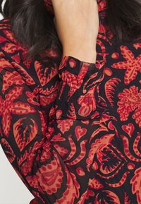 Blusa in tessuto nero trasparente con stampa floreale rossa e arancione. Presenta polsini arricciati con elastico e intricati motivi a foglia.