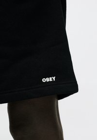 Κάτω μέρος ποδιού με μαύρο σορτς που φέρει μικρό λευκό κεντημένο λογότυπο «OBEY» κοντά στο τελείωμα, πάνω σε ανοιχτό φόντο.