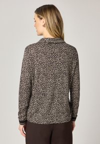 Femme vue de dos portant un haut à manches longues à imprimé léopard avec col et un pantalon sombre, devant un fond uni.