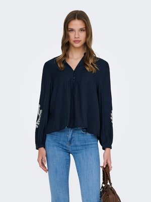 Jonge vrouw draagt een marineblauwe blouse met witte borduursels op de mouwen, lichtblauwe spijkerbroek, en houdt een bruine handtas vast tegen een effen achtergrond.