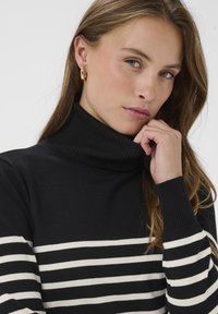 Schwarzer Rollkragenpullover mit weißen horizontalen Streifen, gerippter Textur und enganliegenden Ärmeln. Nahaufnahme eines Modells mit langen Haaren, Hand am Kinn.