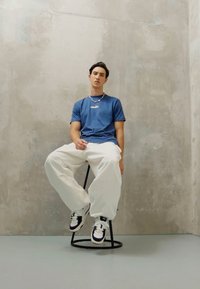 Model i en blå t-shirt, hvide brede bukser og sneakers. Han sidder på en sort skammel op ad en struktureret grå væg. Tilbehør inkluderer en halskæde.