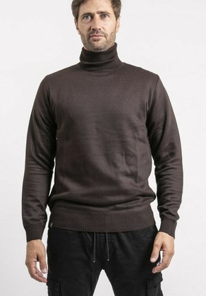 Homme aux cheveux bruns courts portant un pull à col roulé marron foncé et un pantalon noir, sur un fond clair uni.