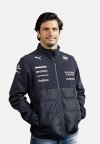 Granatowa kurtka puchowa z zamkiem z przodu, wysokim kołnierzem i akcentami z logo. Wykonana z gładkiego materiału z pikowanymi sekcjami. Noszona z dżinsami.