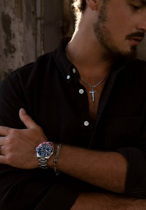 Silberne Metallarmbanduhr mit einer blauen und roten Lünette, ausgestattet mit einem dunklen Ziffernblatt, getragen an einem gebräunten Arm. Die Person trägt ein schwarzes Hemd mit Knöpfen und eine silberne Halskette.