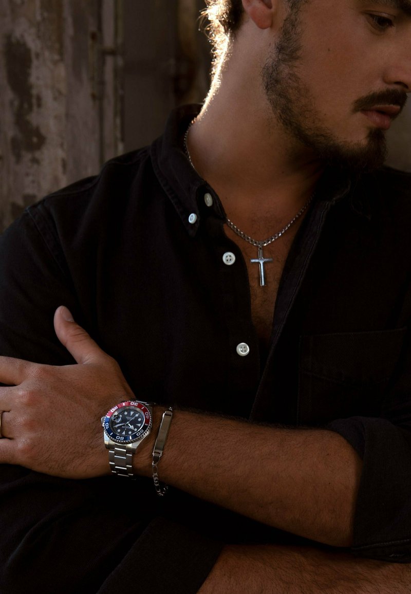 Silberne Metallarmbanduhr mit einer blauen und roten Lünette, ausgestattet mit einem dunklen Ziffernblatt, getragen an einem gebräunten Arm. Die Person trägt ein schwarzes Hemd mit Knöpfen und eine silberne Halskette.