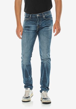 Mann trägt Slim-Fit blaue Jeans, weiße und graue Sneaker, steht vor einem schlichten weißen Hintergrund.