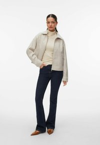 Femme debout portant une veste beige à fermeture éclair, un col roulé crème, un jean bleu foncé et des chaussures pointues marron sur un fond blanc.