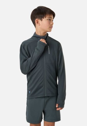 Niño con chaqueta gris oscuro con cremallera y pantalones cortos a juego, mirando hacia un lado, con el pulgar a través del agujero de la manga en una mano.