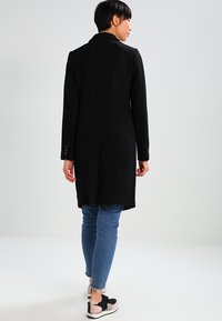 Sort lang jakke med lige snit, der viser knaplukninger på ærmerne og en glat tekstur, parret med lyseblå jeans og sorte sneakers.