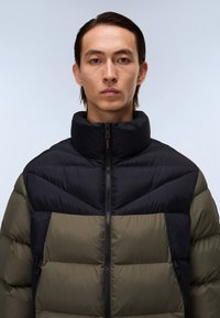 Džemperis pūkų paltas, turintis spalvų blokavimo dizainą juodos ir alyvuogių žalios spalvos, su aukštu apykakle iružtrauktuku, siuvinėtu tekstūros ir šoninėmis kišenėmis.